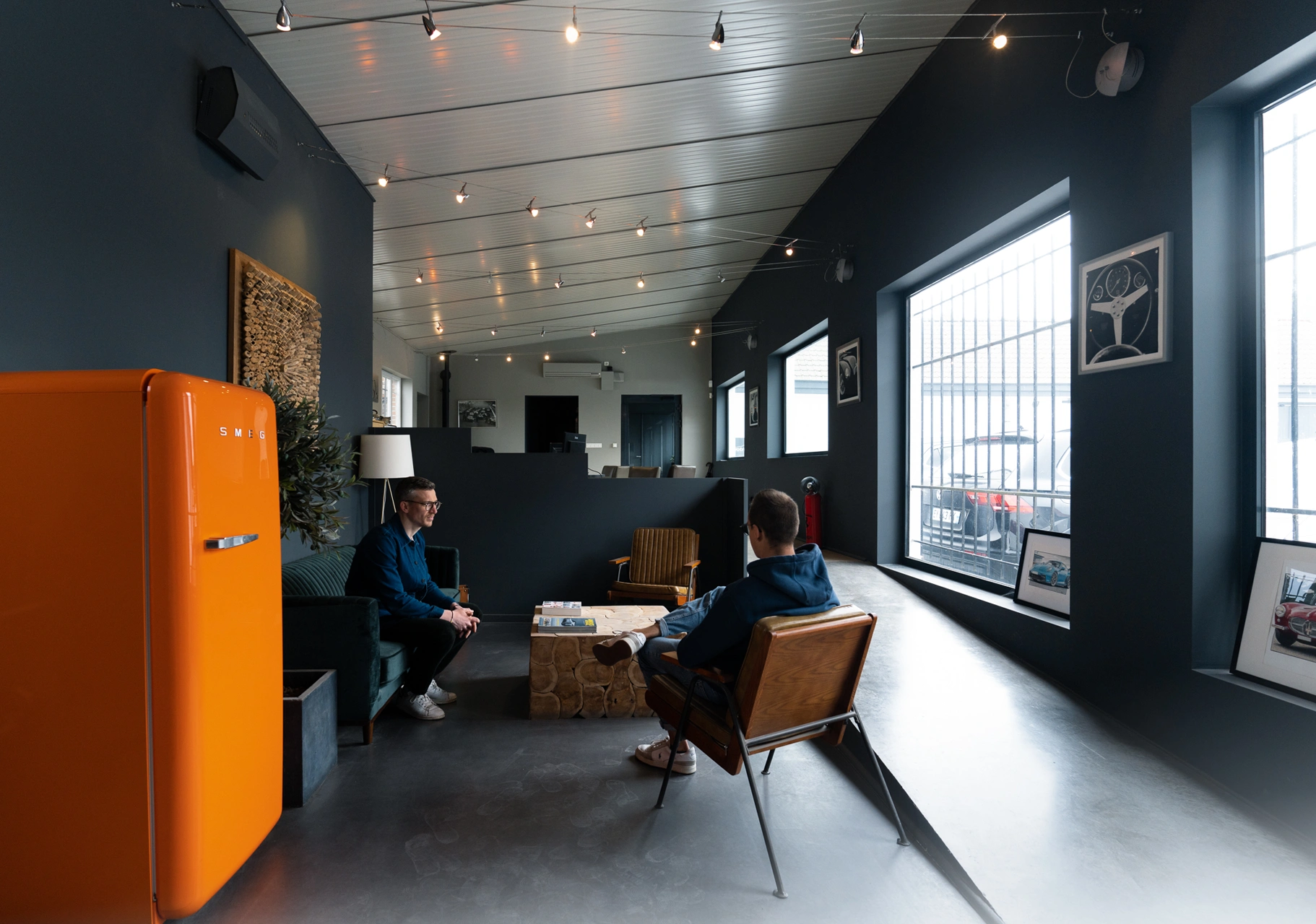 Intérieur moderne avec deux personnes discutant dans un salon, réfrigérateur orange vif et grandes fenêtres.